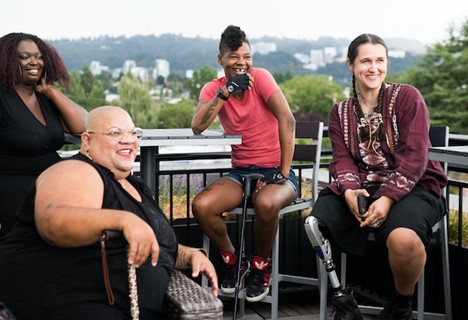 Four disabled people of color with canes and prosthetic legs laugh while chatting. They are on a rooftop deck, in chairs of various height, with greenery and city high-rises in the background.